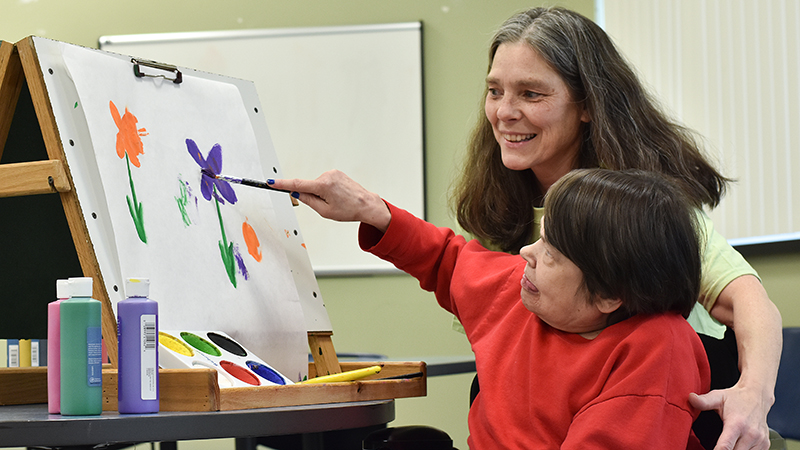 Female employee caregiver helping female client in wheelchair paint flowers on a canvas